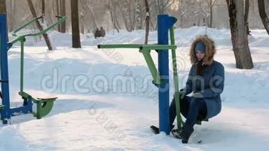 穿着带引擎盖的蓝色羽绒服的年轻女子正在冬季公园的模拟器上做腿部运动。